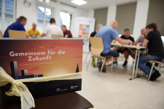 Brettspiel mit dem Titel "Gemeinsam für die Zukunft", im Hintergrund Menschen an Tischen, die spielen.