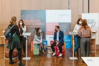 Sechs Menschen vor einem Messestand jeweils in Zweier-Gespräche vertieft. Rechts und links stehen jeweils zwei Personen an Aufstellern, in der Mitte sitzen zwei Personen auf SDG-Würfeln und unterhalten sich.