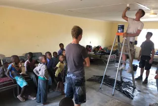Zwei Handwerker arbeiten, einer streicht auf einer Leiter die Decke. Mehrer Grundschul-Kinder schauen ihnen zu.