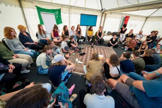 Eine große Gruppe von Menschen sitzt im Kreis in einem Workshop-Zelt.