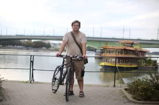 Ein Mann mit Fahrrad steht vor dem Rhein, im Hintergrund ist ein Ausflugsschiff zu sehen.