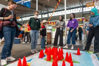 Eine Gruppe von Personen steht in einer Messehalle und schaut auf eine große Weltkarte auf dem Boden. Auf der karte sind rote Pylonen verteilt.