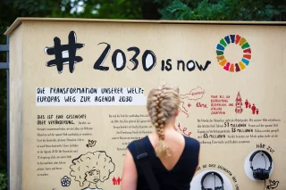 Eine Person steht vor einer Wand, die den Schriftzug "#2030 is now" trägt.
