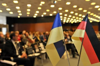 Die ukrainische und deutsche Flagge stehen auf einem Tisch. Im Hintergrund sind Menschen zu sehen