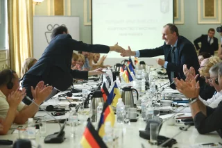 Personen sitzen an einem Konferenztisch, auf dem ukrainische und deutsche Flaggen stehen.