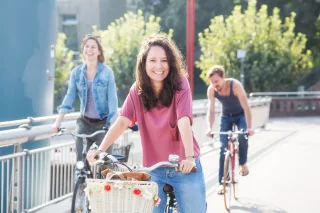 Eine Frau fährt auf einem Fahrrad und blickt lächelnd in die Kamera. Hinter ihr sind zwei weitere Personen auf Fahrrädern. Die Sonne scheint.