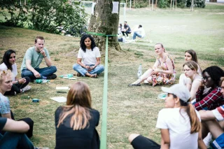 Eine Gruppe junger Menschen sitzt im Kreis, zwischen ihnen ist eine Slackline gespannt. Viele haben Wasserflaschen und Papier vor sich liegen.