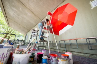 Eine Künstlerin steht auf einer Leiter und bemalt mit roter Farbe eine Wand. Im Vordergrund stehen Farbtöpfe und Pinsel.