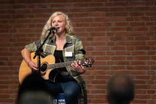 Eine Frau spielt auf einem Hocker sitzend Gitarre und singt.