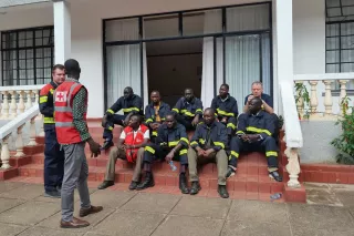 Eine Gruppe von neun Personen, die zum Teil Feuerwehr-Kleidung tragen, sitzen auf einer Treppe. Zwei Personen stehen davor und unterhalten sich.