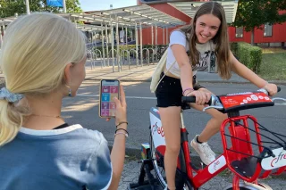 Ein Mädchen hält ein Handy in der Handy. Ein weiteres Mädchen sitzt auf einem Fahrrad.